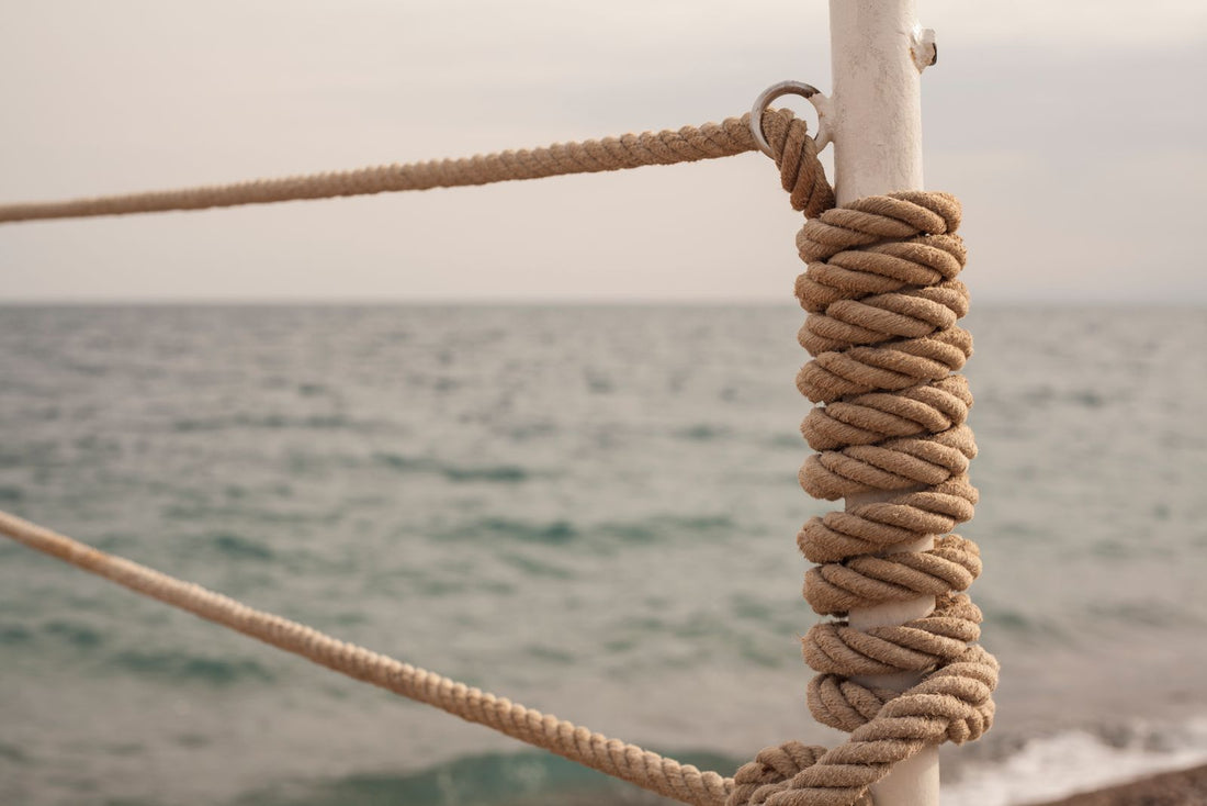 Close-up of a thick rope knotted tightly around a wooden mooring post, with the sea blurred behind it.