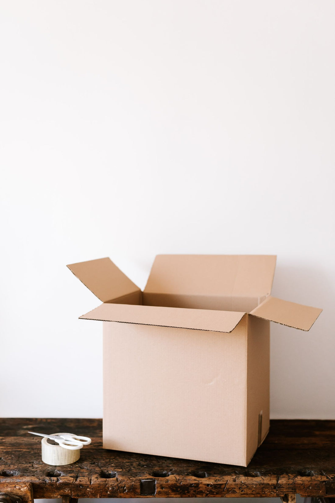 An open cardboard box on a weathered wooden bench with a roll of packing tape and scissors beside it